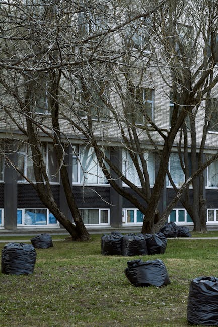 A row of black rubbish bags filled with waste are scattered across a grassy area in front of a modern, multi-storey building with a grey brick façade and large, rectangular windows. The bags are plac