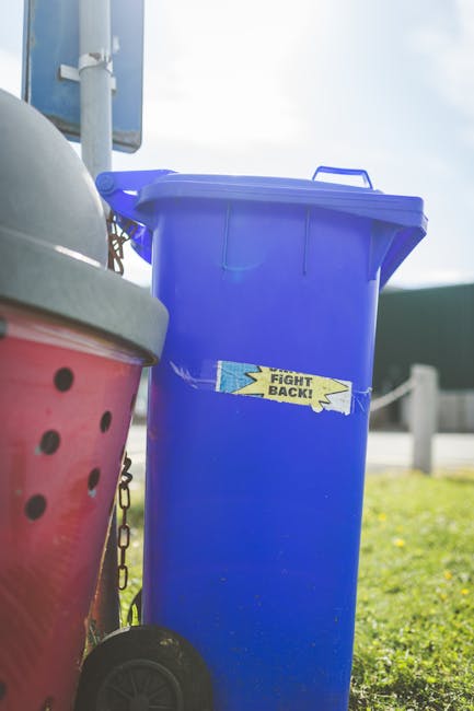A large urban waste collection scene showing multiple overflowing black and black-and-white mixed recycling bins, a bright red refuse bin, and a grey cardboard recycling container, all situated on a p